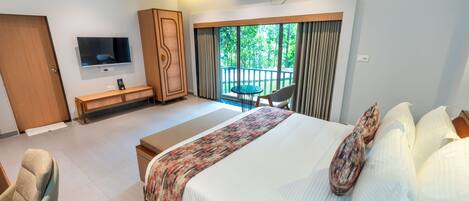 A modern hotel room with a large bed, a TV mounted on the wall, a desk, a chair, and a balcony with a view of greenery.