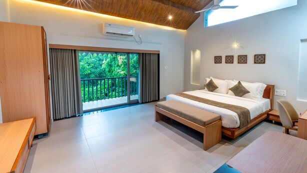 A modern bedroom with a large bed, a desk, and a bench. There is a balcony with curtains and a view of greenery. - Kings Kraft River Side Resort  (Talala)