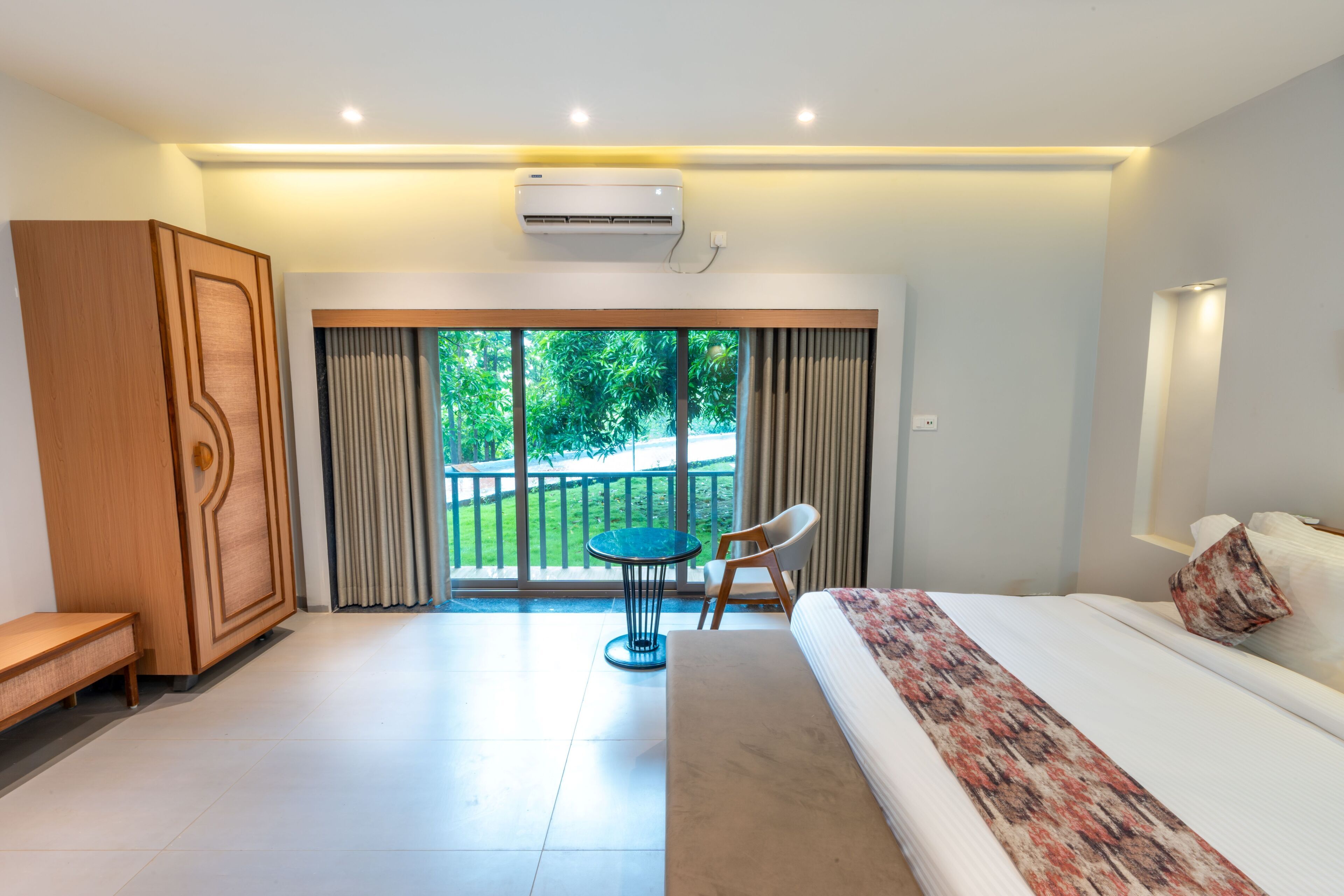 A modern hotel room with a large bed, a wooden wardrobe, a small round table, and a balcony with a view of greenery.