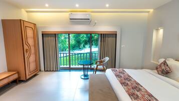 A modern hotel room with a large bed, a wooden wardrobe, a small round table, and a balcony with a view of greenery.
