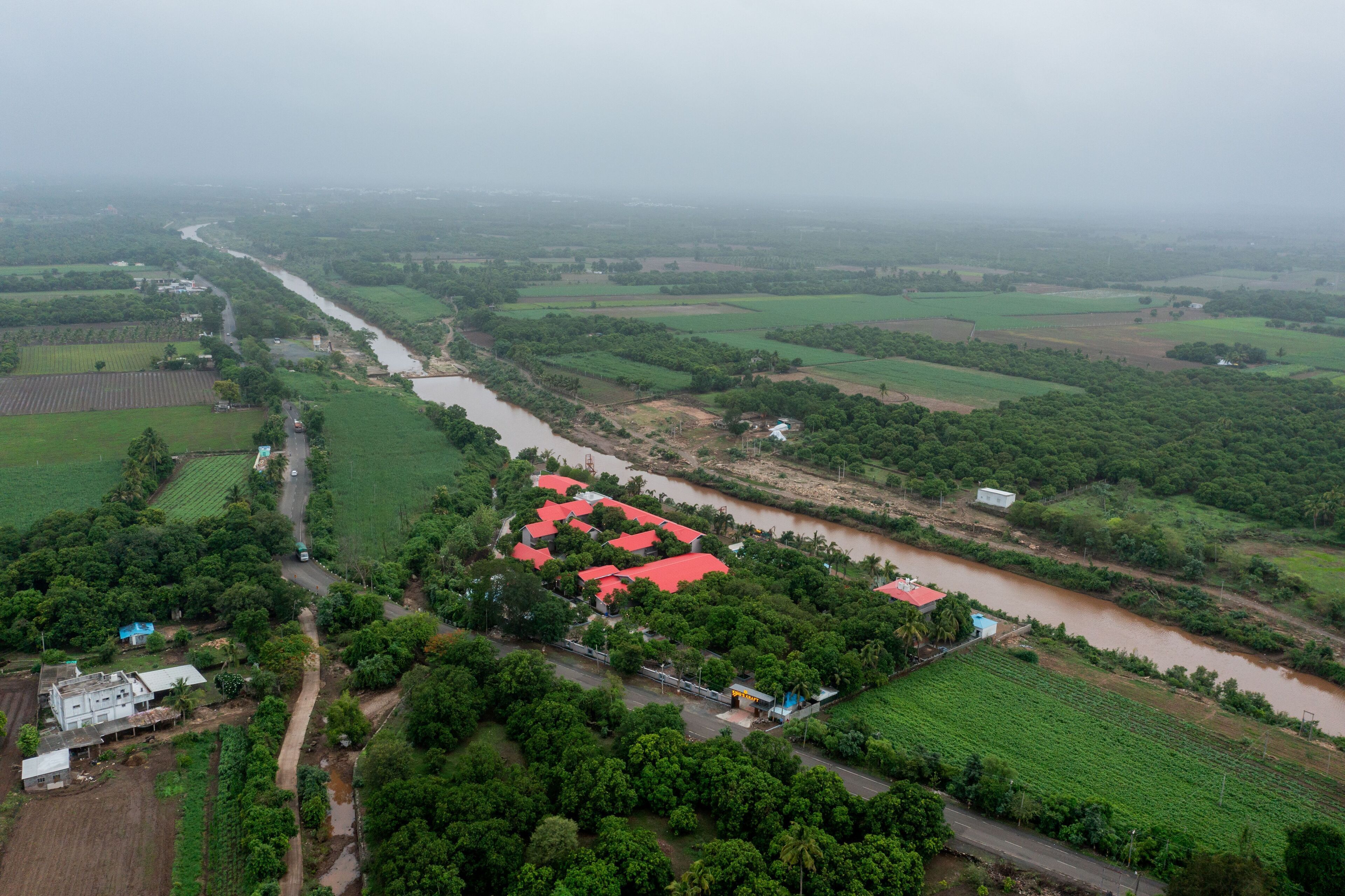 An aerial view of a rural area with fields, a river, and a cluster of buildings with red roofs.
