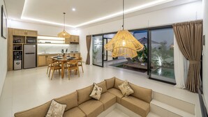 A modern kitchen with wooden cabinets, a dining area with a round table and chairs, a living room with a beige sectional sofa, and large windows with a view of a pool and greenery.