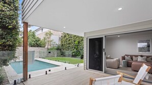 Een modern buitenterras met een zwembad, houten dek en een glazen deur die leidt naar een woonkamer met een bank en kussens.