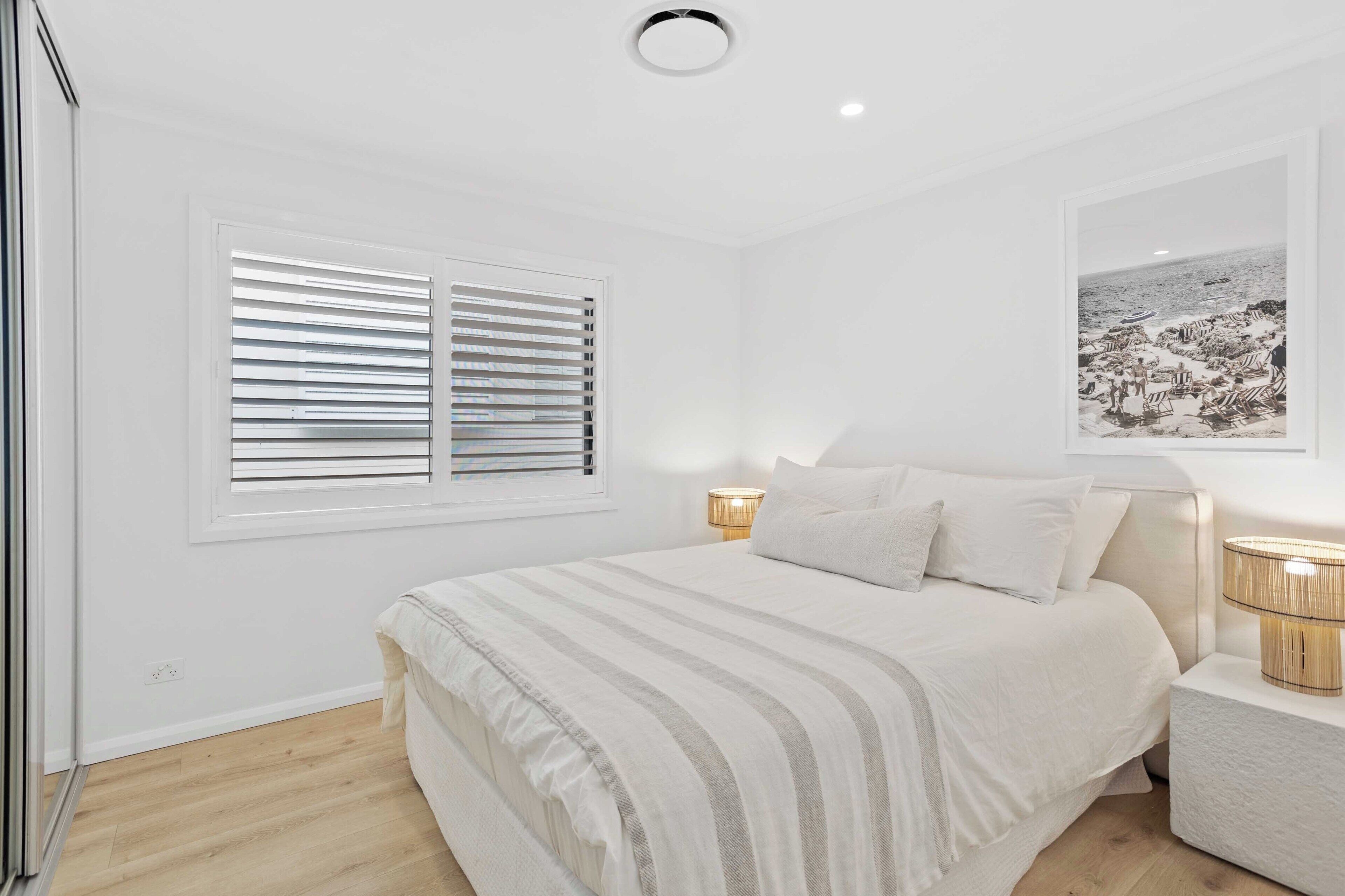 A bedroom with a bed, a window with blinds, a bedside lamp, and a framed picture on the wall.