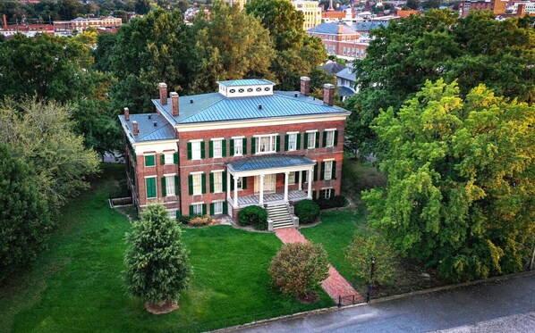 A large, two-story brick building with a blue roof, surrounded by lush greenery and trees.