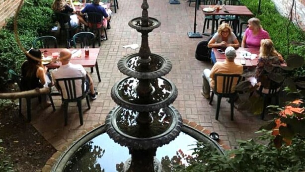 Een terras buiten met tafels en stoelen, een fontein in het midden en een bakstenen muur op de achtergrond.