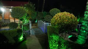 A pathway leading to a building with a red roof, surrounded by green-lit hedges and trees.
