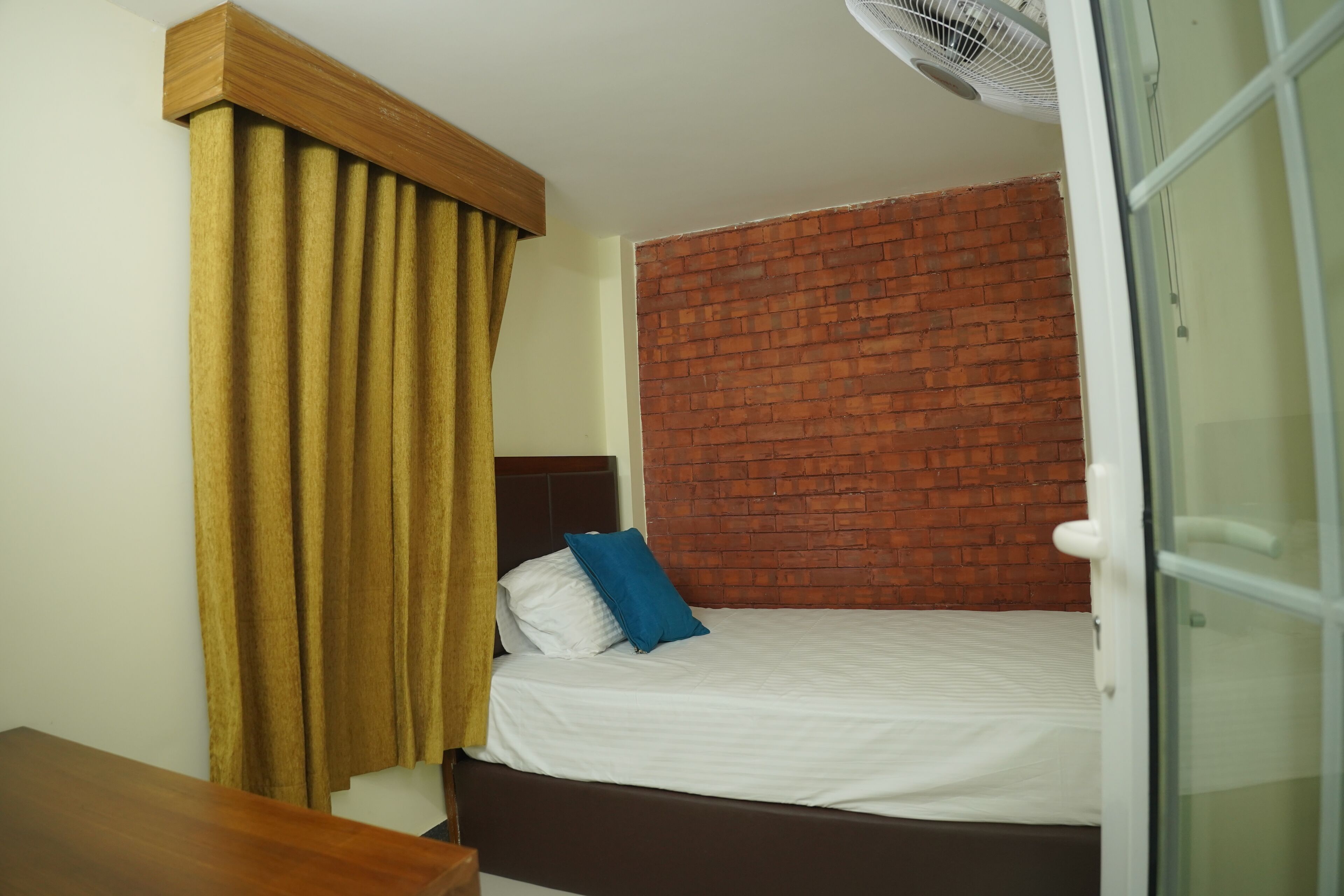 A bedroom with a brick-patterned headboard, a bed with white bedding and a blue pillow, a wooden nightstand, and a window with a curtain.