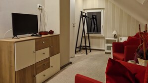 Une pièce de style « loft » avec un plafond en pente, un bureau en bois doté de tiroirs, une télévision, une échelle, une chaise rouge et une fenêtre avec des stores.