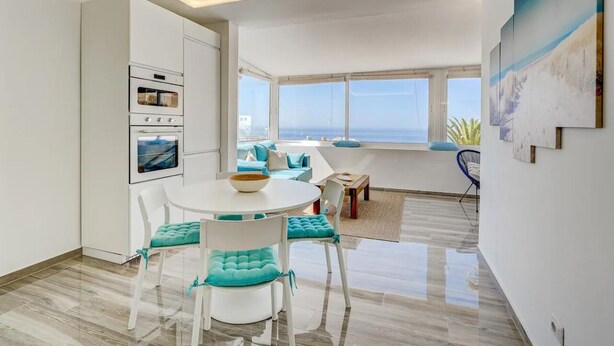 A modern kitchen with built-in appliances, a dining area with a round table and chairs, and a view of the ocean through large windows. - Ocean View Montesol Apartment with 2-bedrooms and outdoor swimming pool (Costa Adeje)