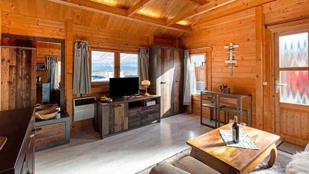 Interior de una cabaña de madera con televisión, muebles de madera, una mesa de comedor y una ventana con vistas.