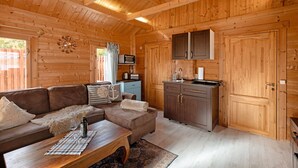 Interior de una cabaña de madera con sofá, reposapiés, mesa de centro de madera, zona de cocina con armarios y un microondas.