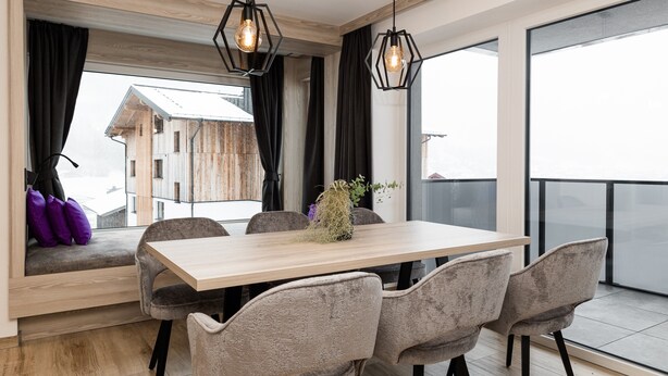 Ein moderner Essbereich mit einem Holztisch, grauen Polsterstühlen und Blick auf ein Holzgebäude durch große Fenster.
