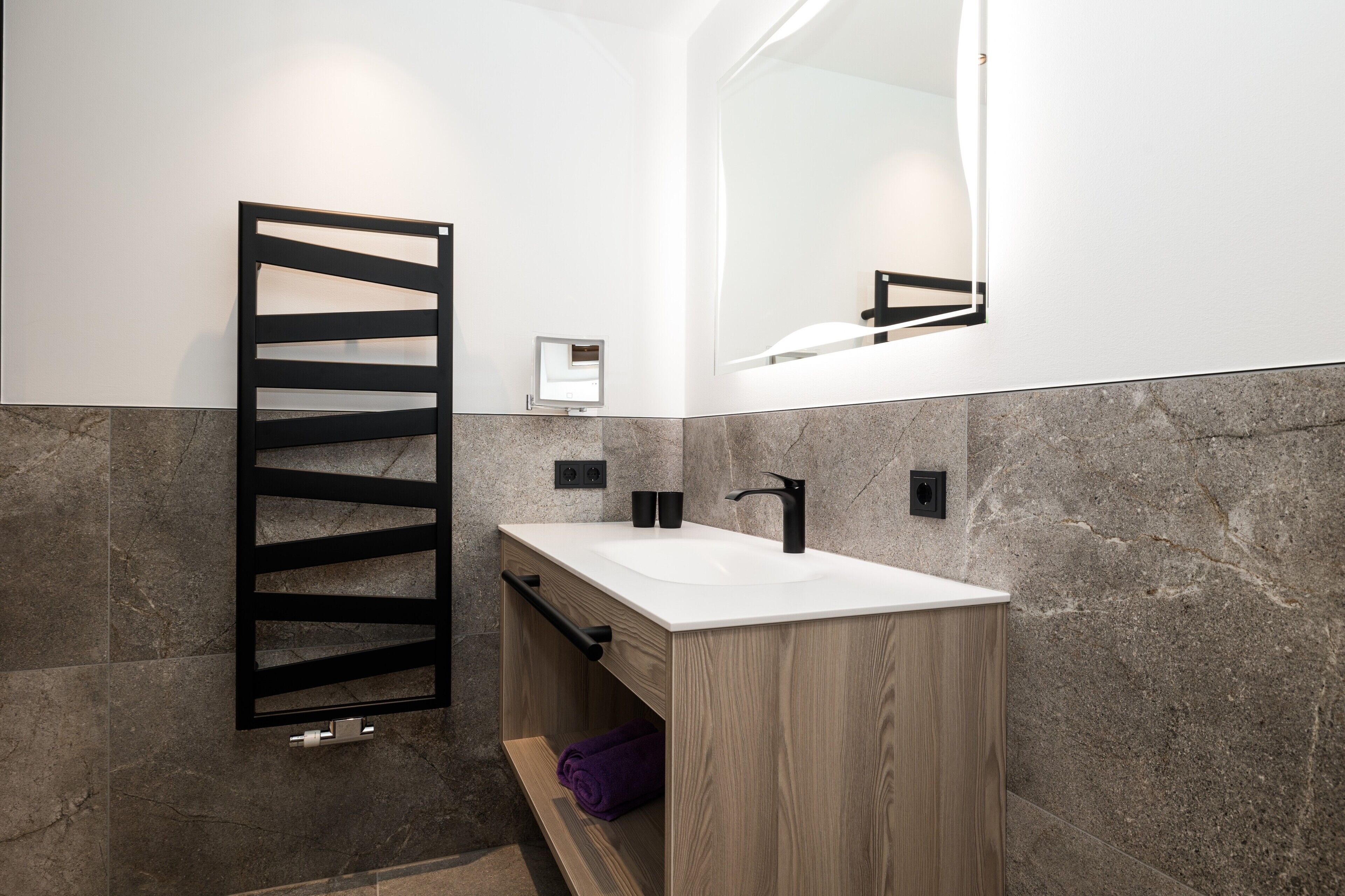 A modern bathroom with a wooden vanity, a black towel rack, a white sink, and a large mirror.