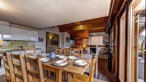 Un comedor con una mesa de madera dispuesta para seis personas, rodeada de sillas de madera. Se ve una cocina con electrodomésticos modernos y una chimenea de piedra al fondo.