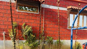 Un columpio con cadenas de metal y asientos negros, un muro de ladrillo rojo y una ventana que da a zonas verdes.