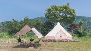 Un campamento con dos tiendas de campaña, una marrón y otra blanca, montado en una zona cubierta de césped con vistas a una colina boscosa.