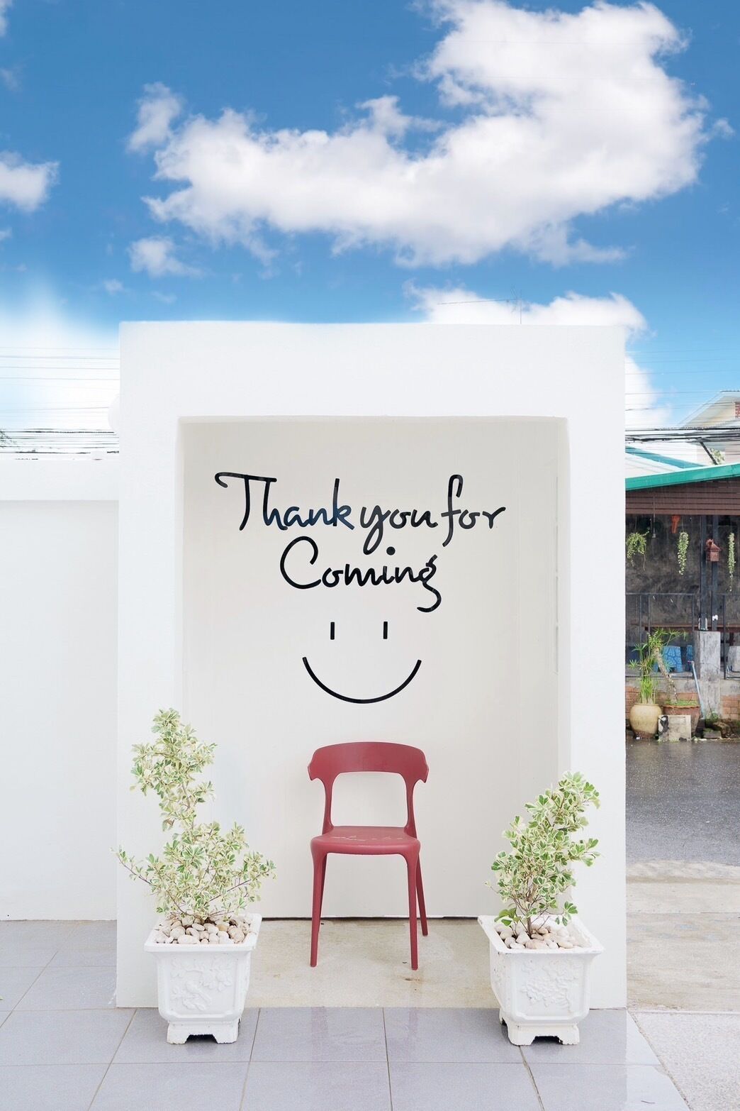 Un letrero que dice 'Thank you for Coming' con un carita sonriente, una silla roja y dos plantas en macetas.