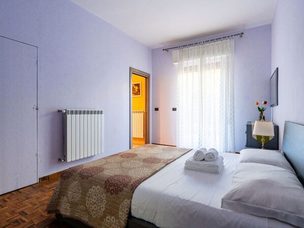A hotel room with a bed, a radiator, a window with curtains, a nightstand with a lamp, and a door leading to another room.