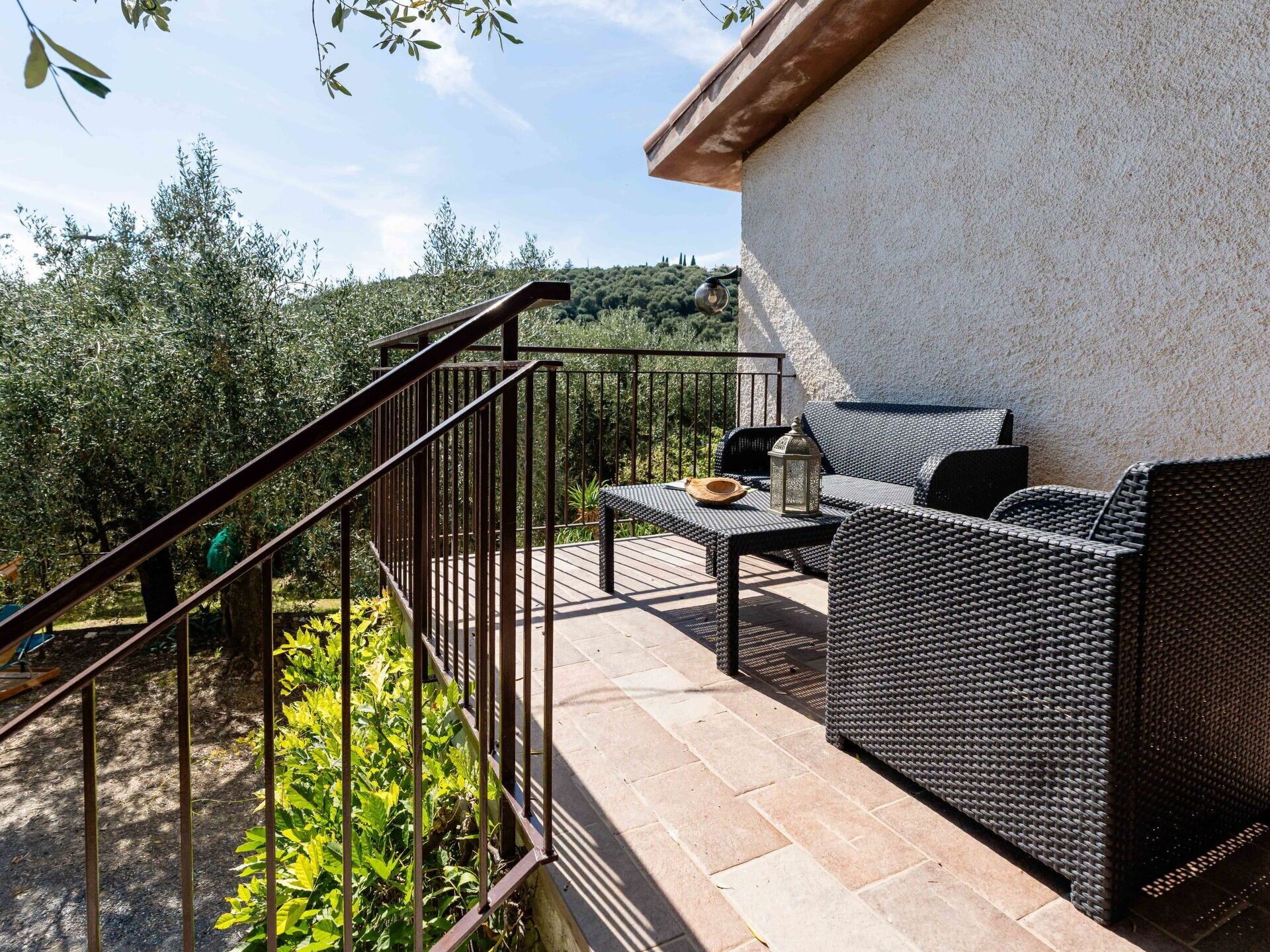 Een terras buiten met rieten meubilair, een metalen hekwerk en uitzicht op groen en heuvels.