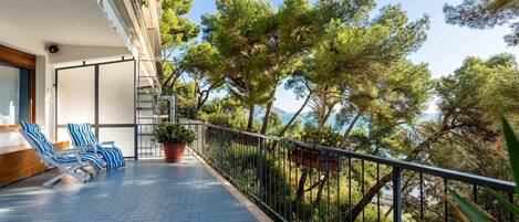 A balcony with blue striped chairs, a potted plant, and a view of trees and the sea.