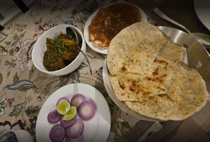 Ein Tisch mit einer blumigen Tischdecke, ein Teller mit gehackten Zwiebeln, eine Schüssel Curry, ein Teller mit Fladenbrot und eine Metalltablett mit mehr Fladenbrot.