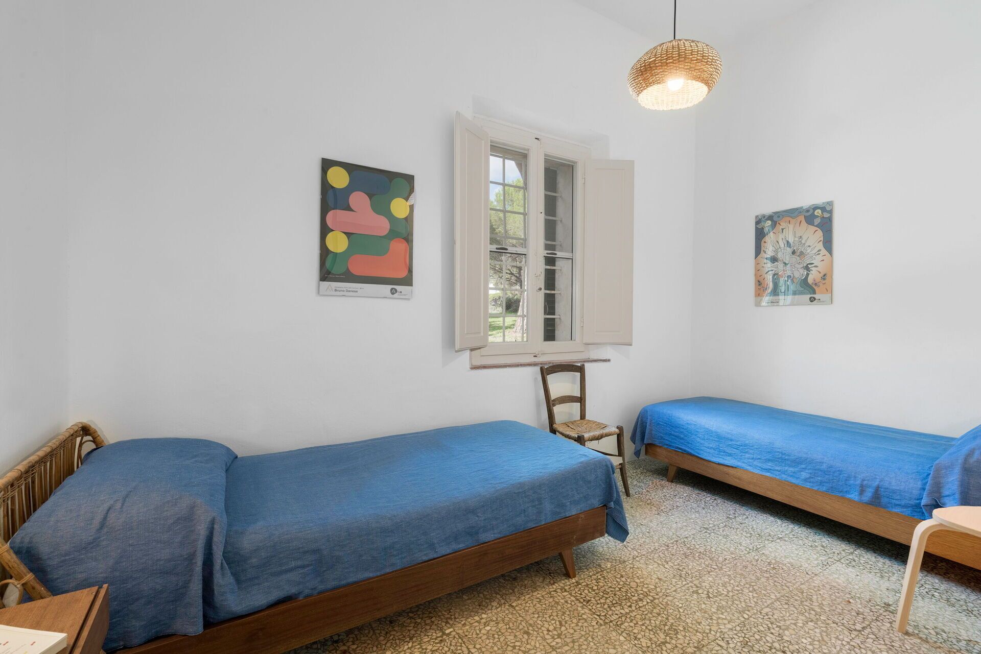 Une chambre avec deux lits simples, une fenêtre dont les volets sont ouverts et deux œuvres d’art accrochées au mur.