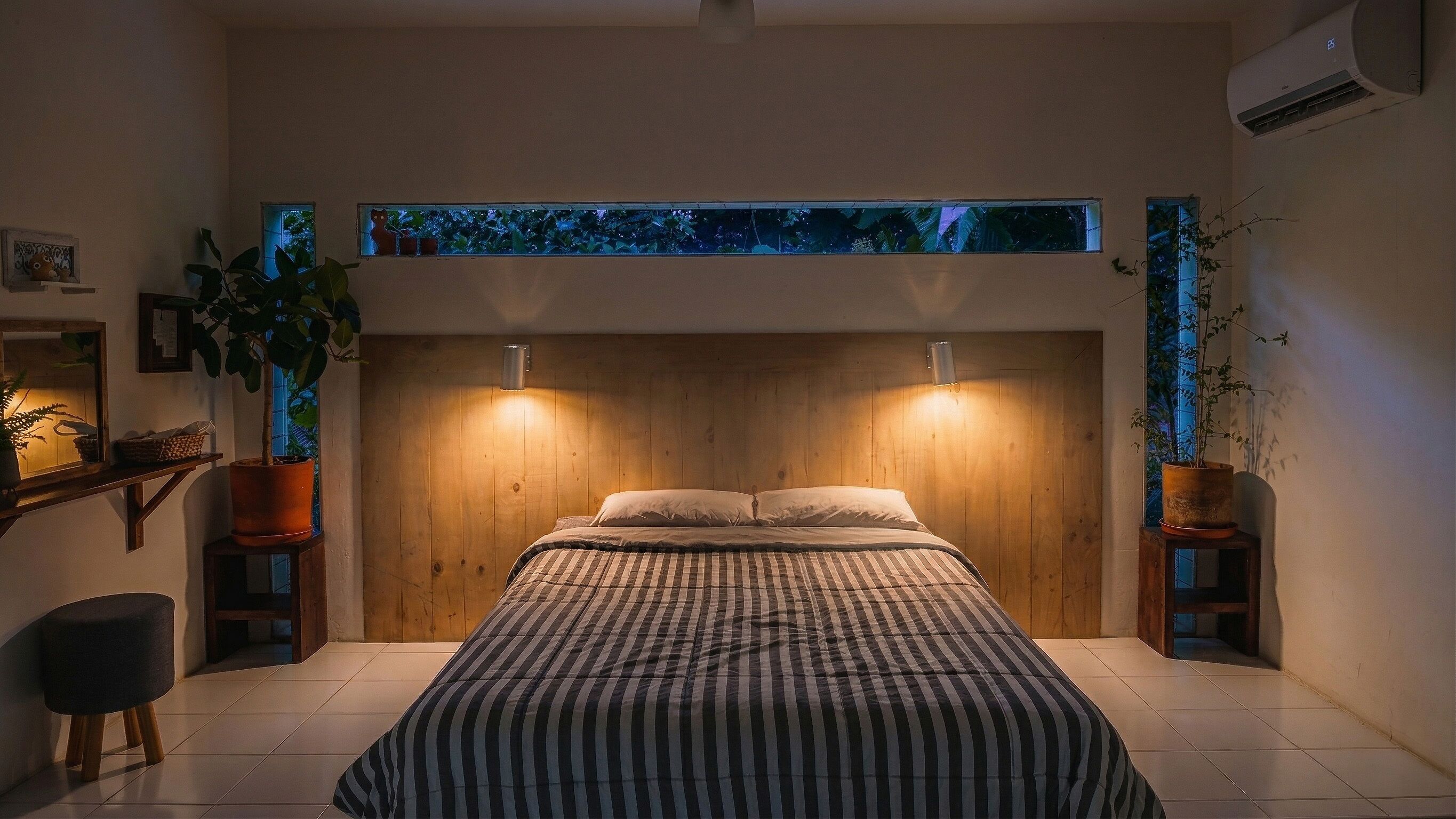 Een slaapkamer met een houten hoofdeinde, een bed met gestreept beddengoed, twee wandlampen en een uitzicht op de lucht door een raam.
