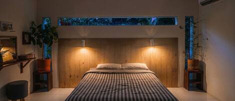 Een slaapkamer met een houten hoofdeinde, een bed met gestreept beddengoed, twee wandlampen en een uitzicht op de lucht door een raam.