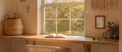 A room with a wooden desk, a wicker chair, a window with a view of greenery, and a macrame wall hanging.