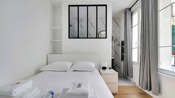 Une chambre d’hôtel avec un lit, du linge de lit blanc, une table de chevet, une lampe et une fenêtre avec des rideaux.