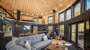 A spacious interior with a large gray sofa, a wooden staircase, and large windows offering views of the outdoors.