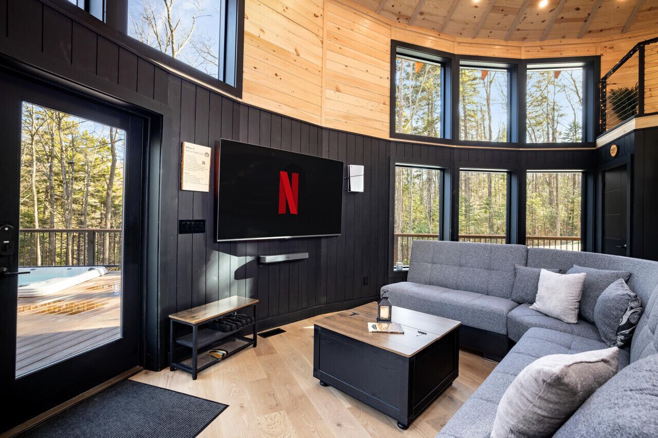 Una sala de estar moderna con un televisor de pantalla plana grande montado en la pared, un sofá seccional gris, una mesa de centro y una terraza de madera visible a través de la puerta de vidrio.