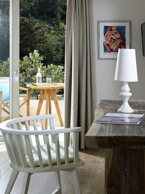 Enjoy a serene view of the lush landscape from the comfort of this stylish room, featuring a glass door, a wooden table, a chair, a lamp, and a beautiful abstract painting.