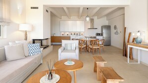 Ein modernes Wohnzimmer mit Sofa, Couchtisch und Sesseln. Im Hintergrund ist eine Küche mit Schränken und einem Kühlschrank zu sehen.