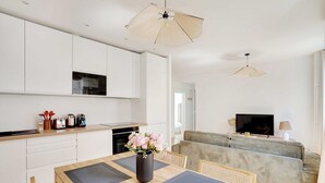 Una cocina moderna con armarios blancos, una mesa de comedor con individuales, un florero con flores y un televisor en la sala.