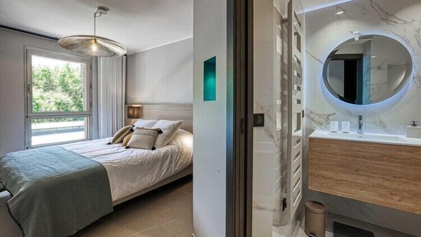 Une chambre moderne avec un lit, une grande fenêtre, une salle de bain dotée d’un meuble-lavabo en marbre et d’un miroir rond.