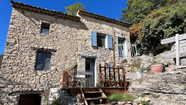 A stone building with a wooden staircase leading to a door, surrounded by rocks and vegetation. - Domaine du Cruvelet (Trigance)