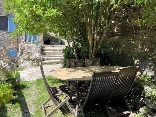 An outdoor dining area with a round table and metal chairs, surrounded by greenery and a stone wall.