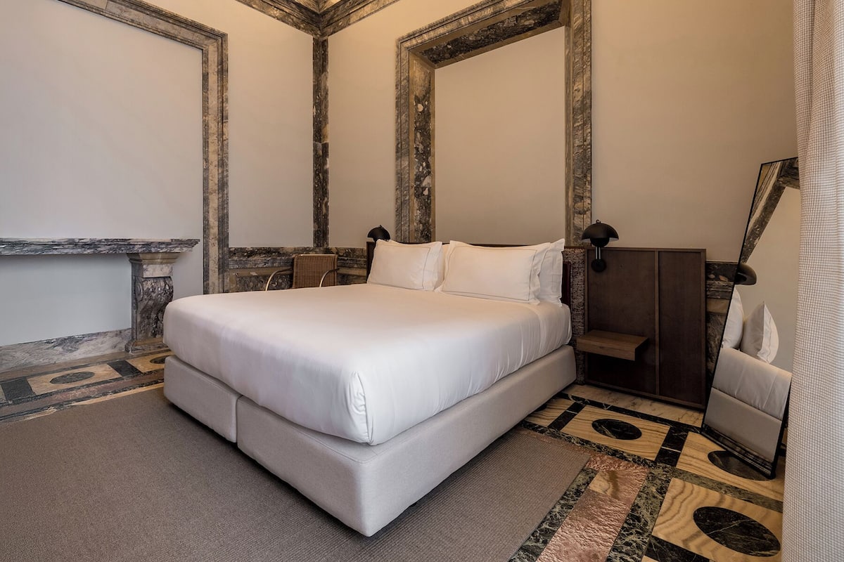 Quarto de hotel com uma cama grande, uma mesinha de cabeceira de madeira, uma cadeira e um espelho.