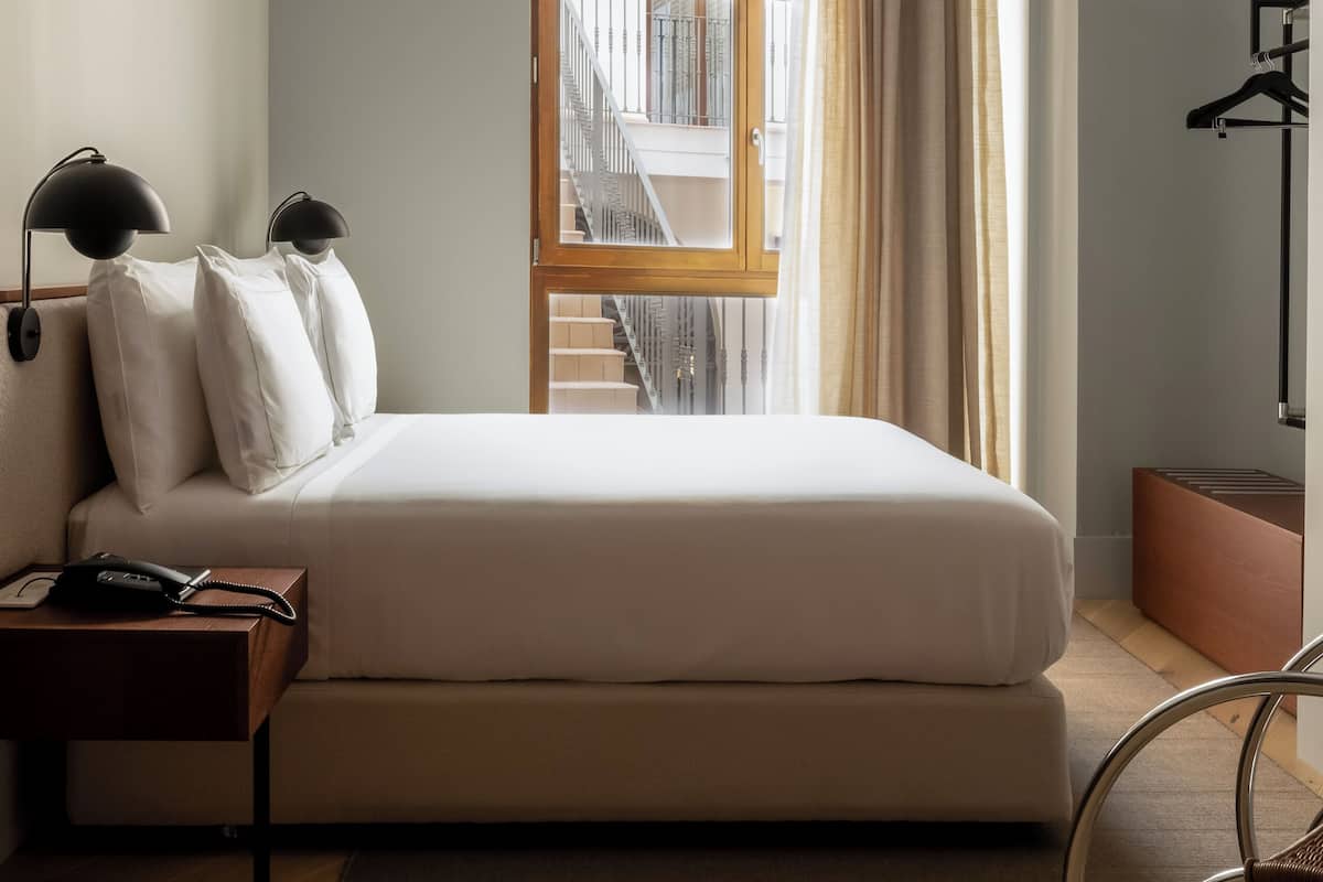 Quarto com uma cama, mesa de cabeceira, abajur e vista para uma escadaria através de uma janela em arco.