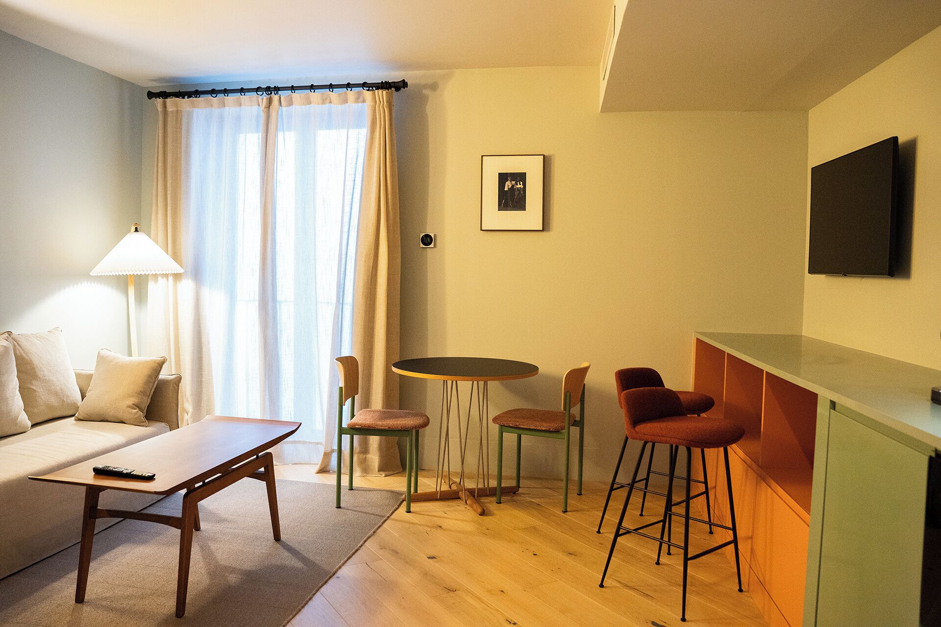 Ein Wohnzimmer mit einer Couch, einem Couchtisch, einem Esstisch mit Stühlen, einer Lampe und einem an der Wand befestigten Fernseher.