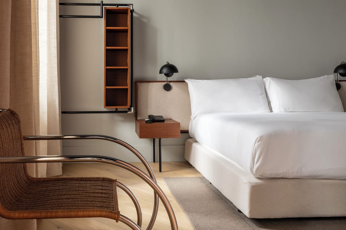 Quarto de hotel com cama, mesa de cabeceira, luminária, cadeira de vime e estante de madeira.