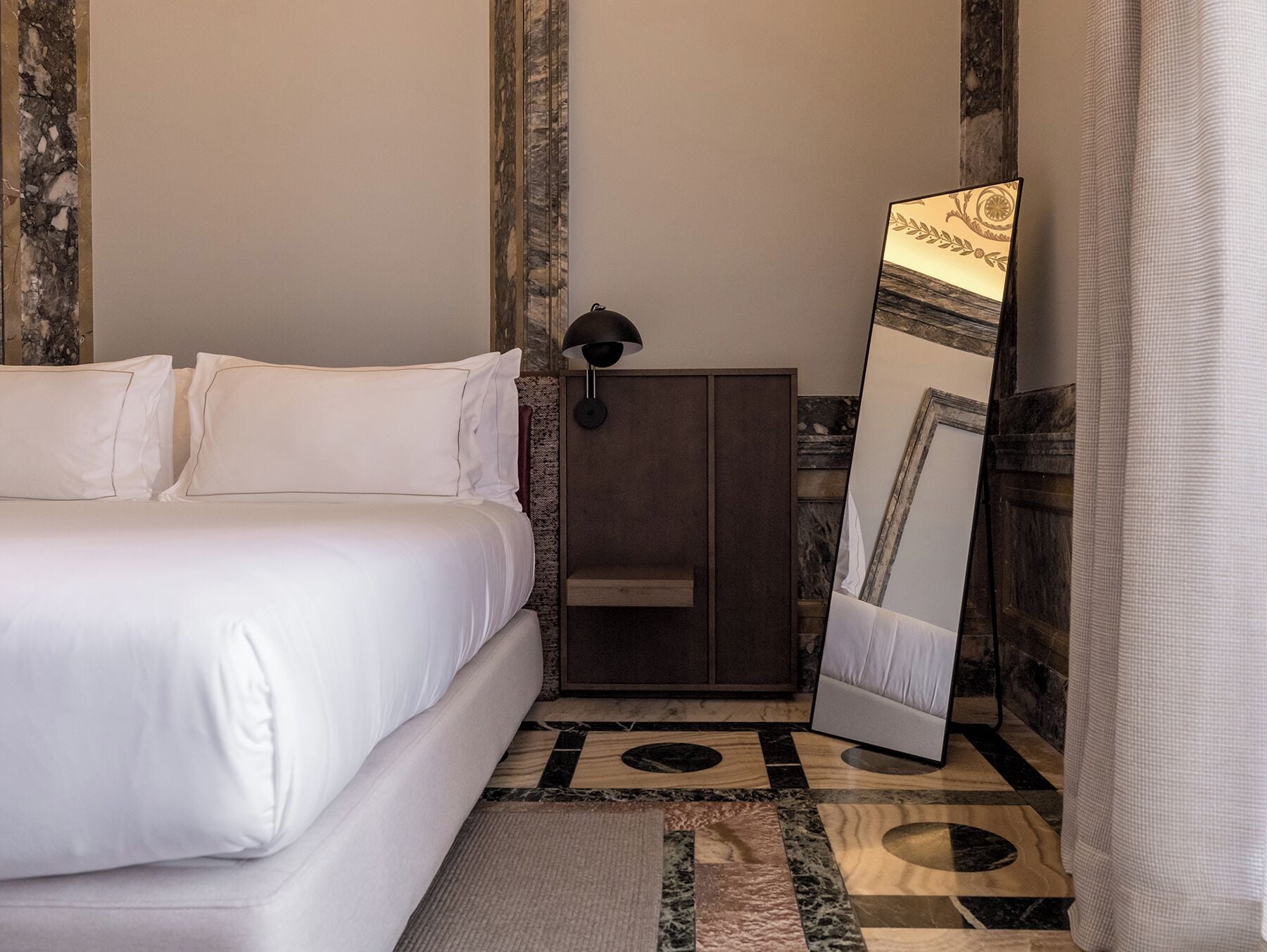 Un lit bien fait, avec du linge de lit blanc, une armoire en bois, un grand miroir doté d’un cadre décoratif et un sol à motifs.