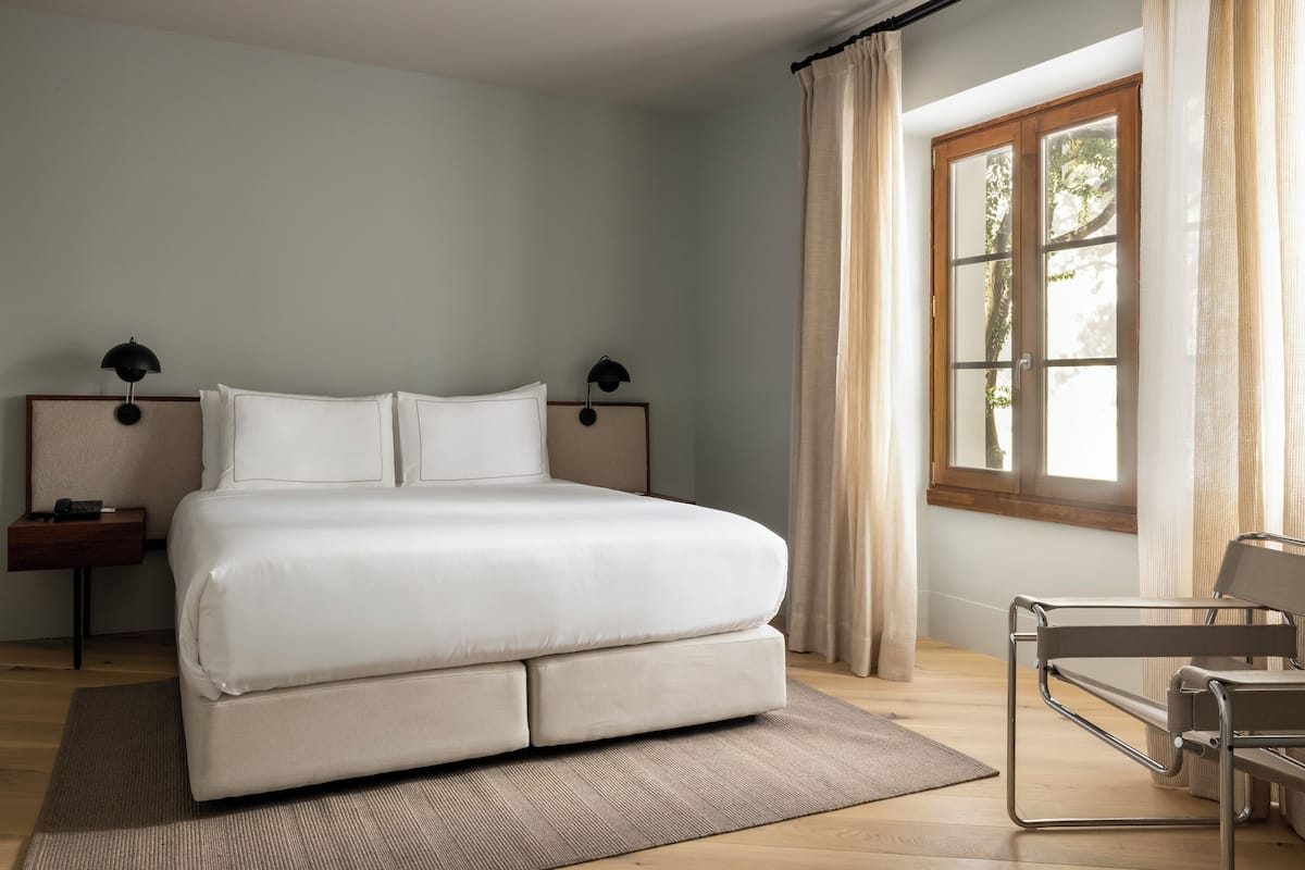 Um quarto moderno com uma cama grande, uma mesinha de cabeceira, uma cadeira e uma janela com cortinas.