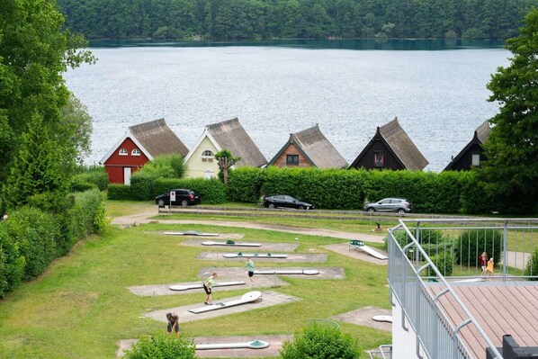 A lakeside view with thatched-roof cottages, a grassy area with exercise equipment, and a red building.