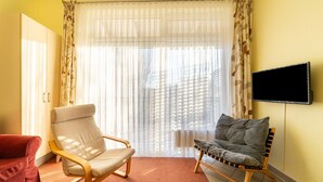 Une pièce comprenant un fauteuil beige, un canapé rouge, une chaise en bois et un téléviseur fixé au mur.
