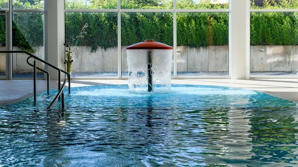 Uma piscina com uma fonte de água, rodeada por grandes janelas e um muro de concreto.