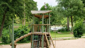 A wooden play structure with a rope ladder and a platform, surrounded by sand and greenery.