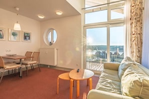 Een moderne woonkamer met een bank, een eettafel met stoelen, een ronde tafel met een afstandsbediening, een groot raam met uitzicht, en een radiator.